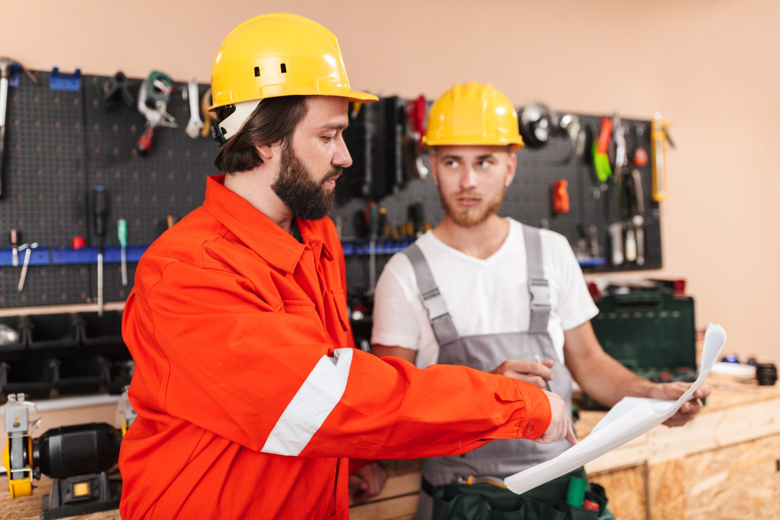 Two builders in work clothes and hardhats working in workshop wi Two builders in work clothes and yellow hardhats working in workshop with tools on background